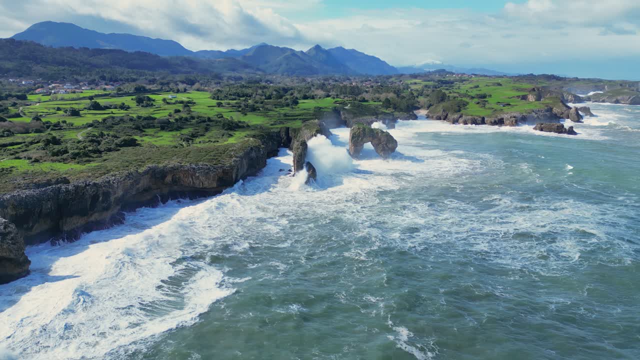Powerful ocean waves crash against a rugged cliffside, rushing through a rock arch. Majestic mountains rise in the distance, creating a dramatic and wild seascape that highlights the power of nature.