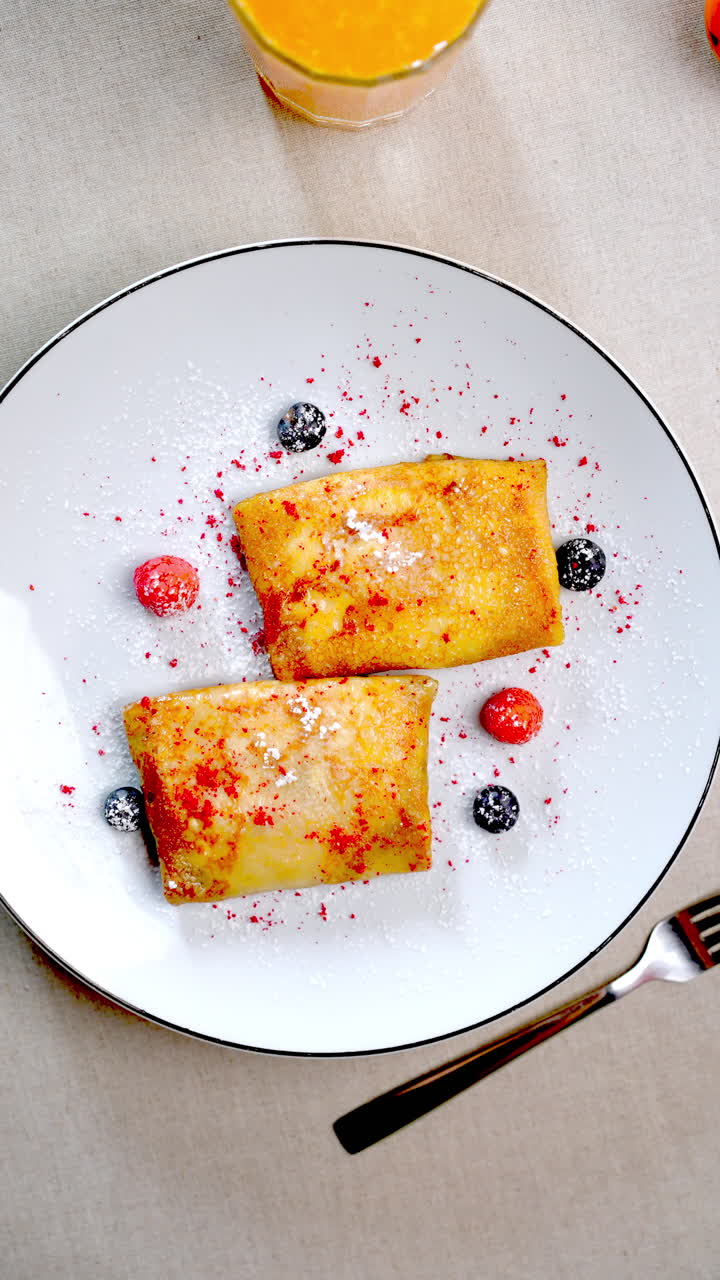 Stop motion animation revealing nalysnyky, traditional Eastern European crepes garnished with berries and powdered sugar, served on white plate with orange juice