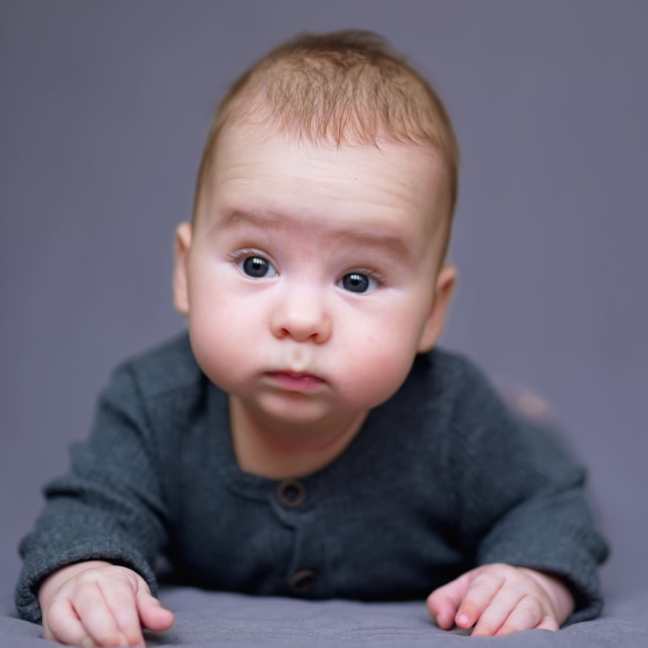 Adorable little child in grey clothes lies on belly in front of camera. Sweet funny boy with plump cheeks making faces. Close up