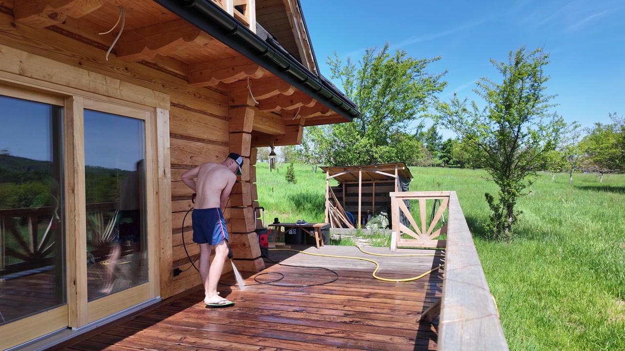 Rear view of a man standing on a wooden deck in front of a traditional log cabin with large glass doors and an empty firewood shelter.