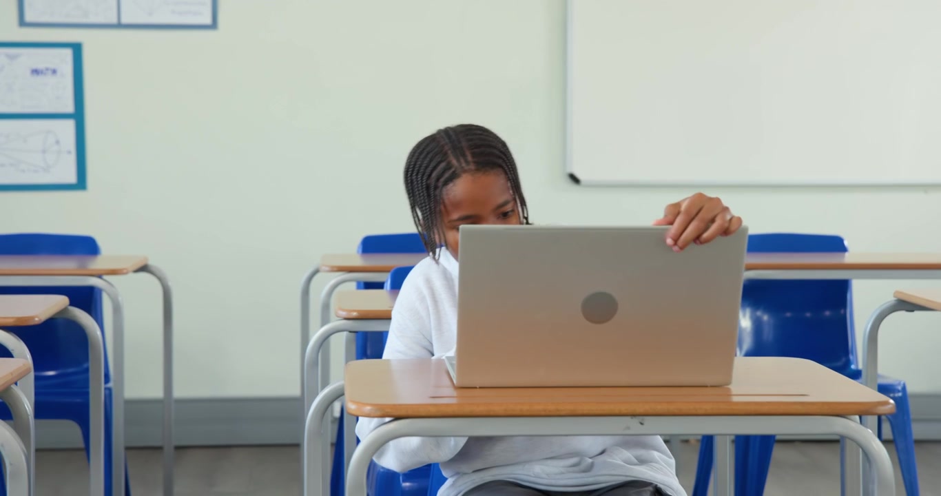 Adolescente maschio africano con felpa grigia con cappuccio che trova un banco di scuola vuoto, posa lo zaino e apre il laptop per studiare.