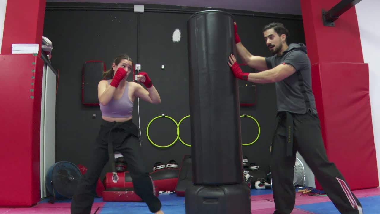 Woman trains boxing and kicks on boxing training device - man helps and holds against it