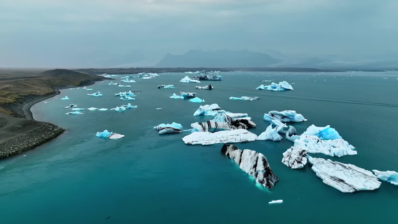 A smooth aerial orbit reveals drifting Icelandic icebergs scattered across a wide lagoon, with layered blue surfaces, dark streaks, and a rugged shoreline under a cold Arctic sky