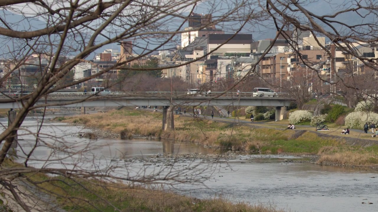Static wide shot of a riverbank, bridge, and urban buildings under a clear winter sky.