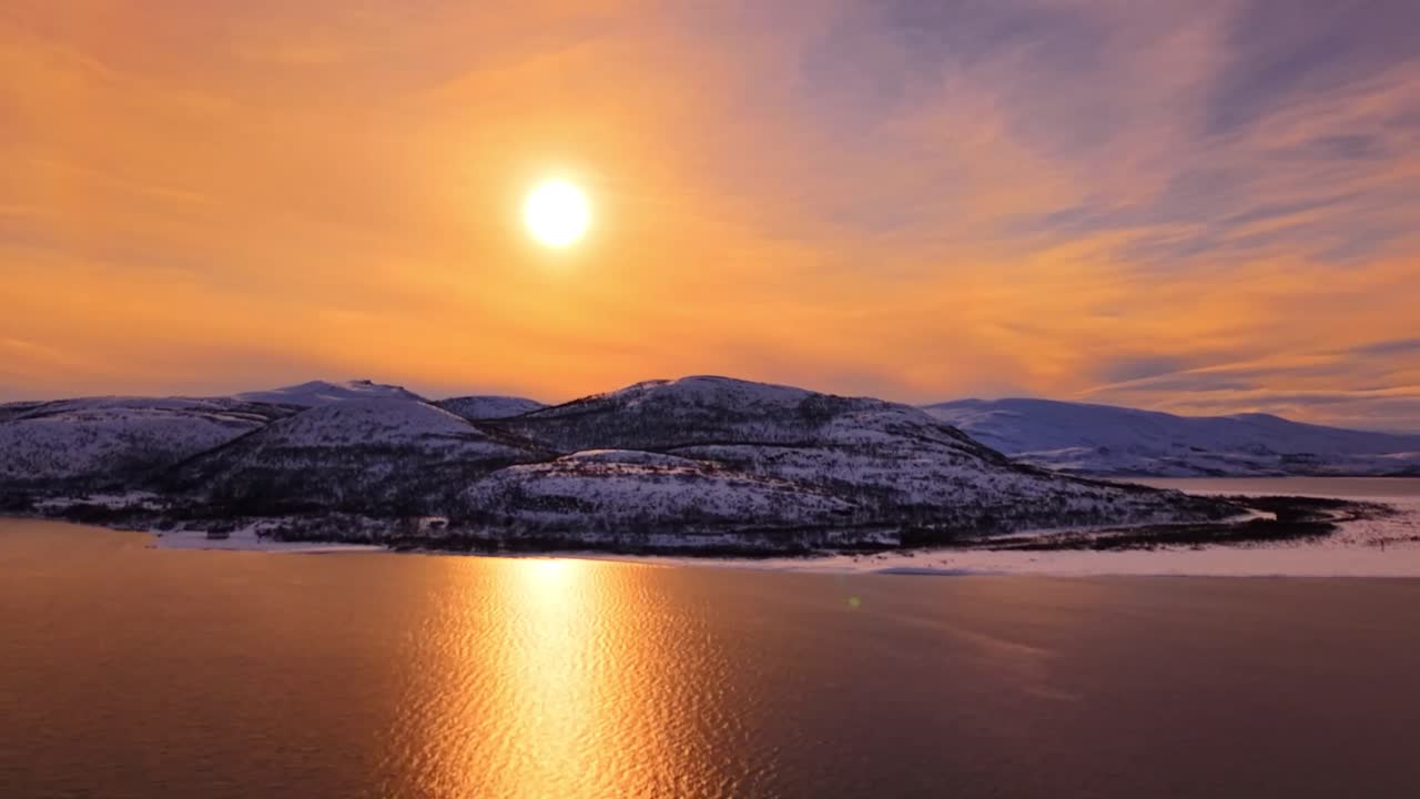 Aerial drone footage gliding and flying over Bartens sea golden reflective water and is large Giemaš white snow covered mountains is in the background with a large orange sun during sunset or sunrise.