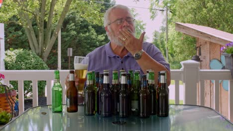 an elderly man sits at a table in his backyard, sampling different beers. he examines each bottle and enjoys the process of tasting, surrounded by various beer bottles