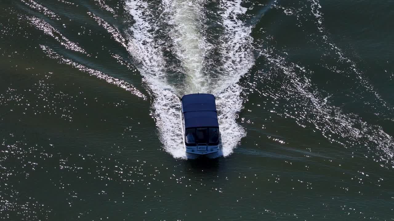 Drone follows houseboat moving steadily along sunlit canal, creating ripples in calm water