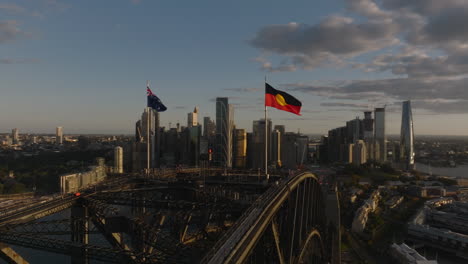 Australian-and-Aboriginal-Flag-on-Top-of-Sydney-Harbor-Bridge,-Golden-Hour-Sunset,-With-Sydney-City-Skyline-in-Background,-Aerial