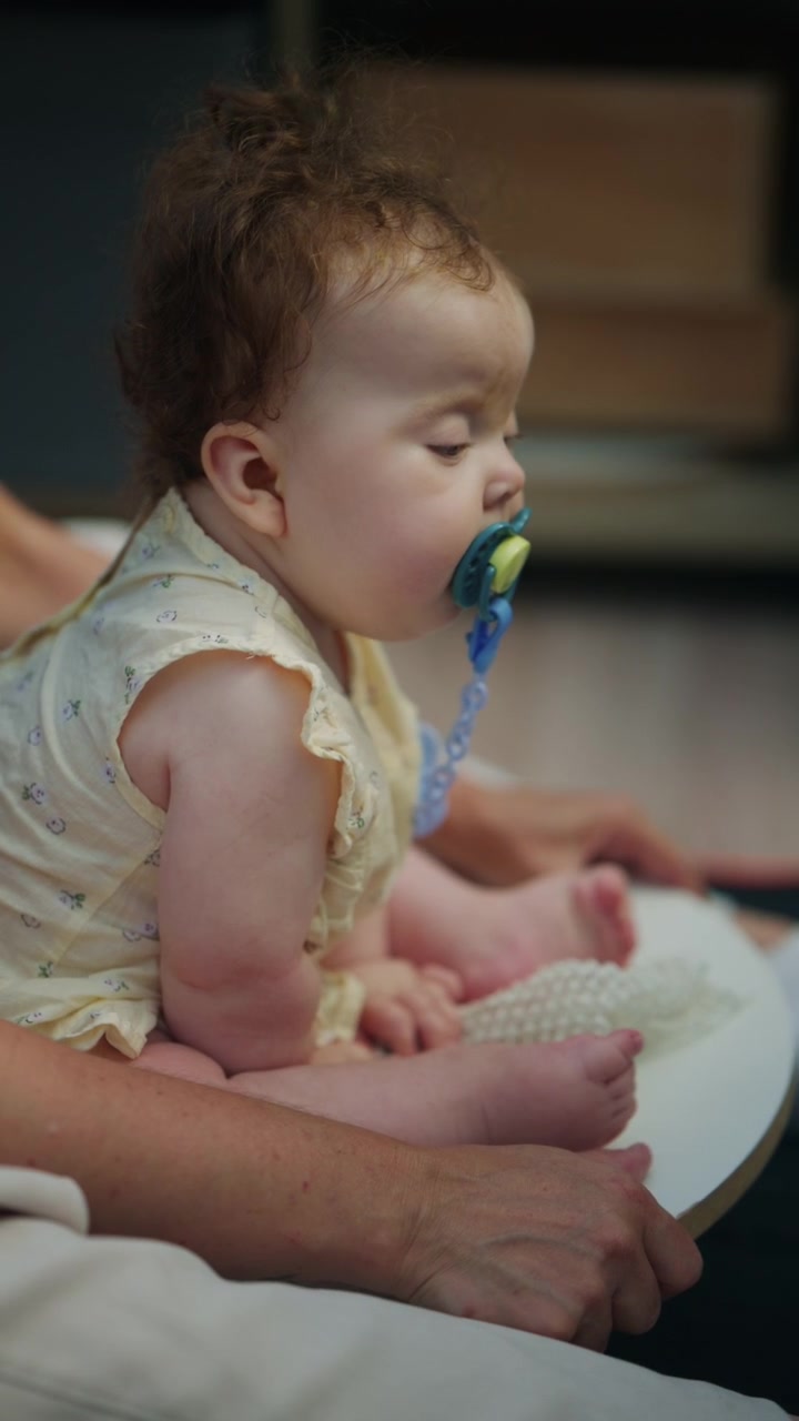 Baby sitting with adult support on a soft indoor mat, wearing a light yellow outfit during a calm caregiving moment