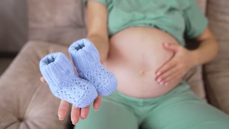 Expectant mother holding pretty blue baby socks on her palm. Mother caressing her big belly lovingly. Knitted tiny socks for newborn boy close up.