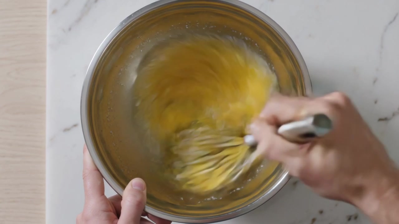 Mani che sbattono e mescolano uova rotte in una ciotola d'acciaio su un bancone di marmo, preparando uova sbattute con una frusta.