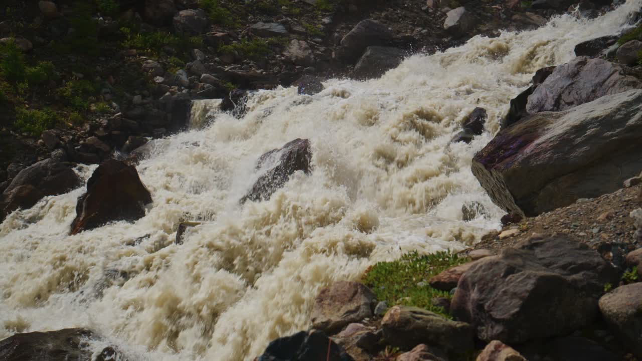 Fiume di montagna impetuoso si infrange sulle rocce, agitando acqua bianca. Il flusso turbolento schizza contro pietre frastagliate. Muschio verde si aggrappa a massi umidi accanto alla corrente impetuosa