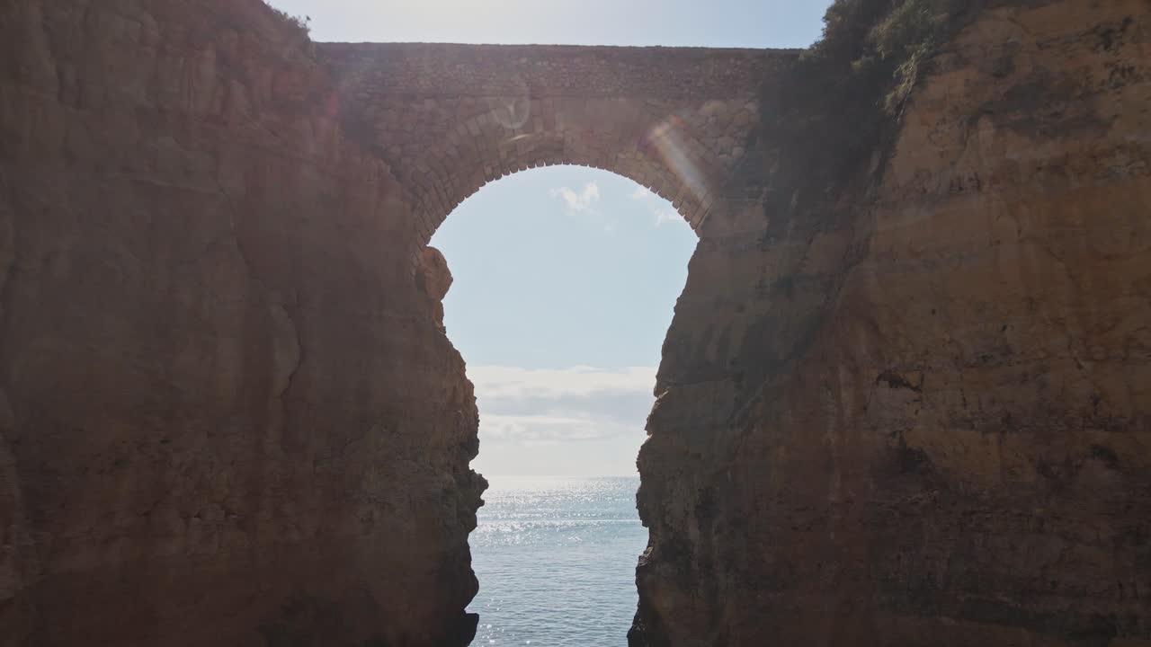 Aerial drone flies forward, passes through the iconic stone bridge, and continues across towering cliffs revealing the Algarve coastline and sparkling sea beyond in Lagos, Portugal