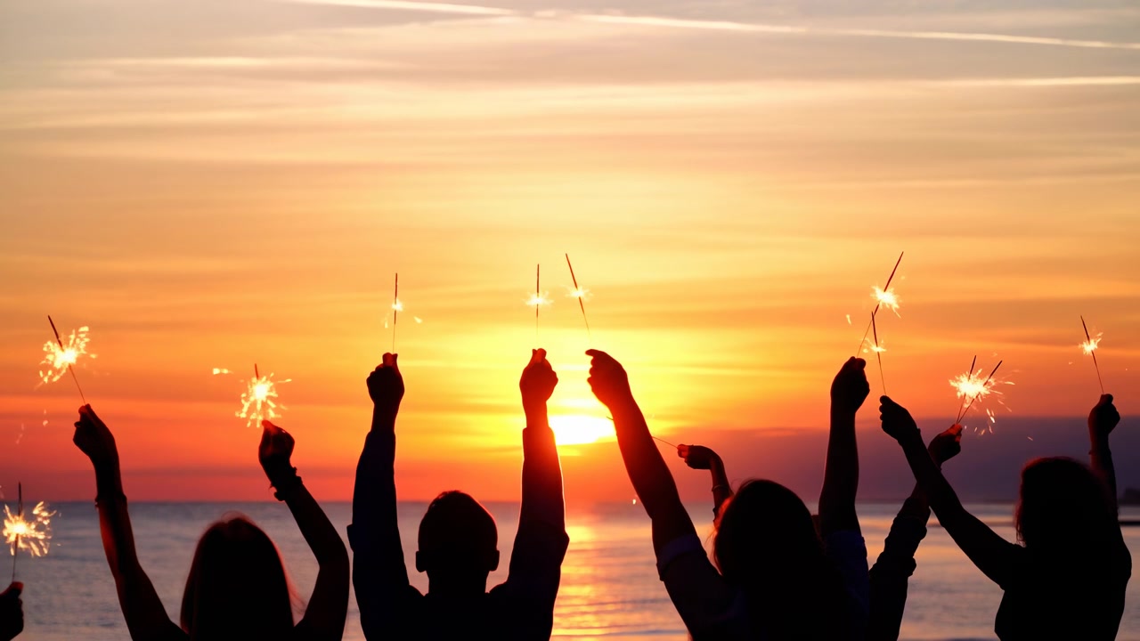 Die Sequenz zeigt eine Gruppenfeier, die vom frühen Sonnenuntergang bis in die Dämmerung fortschreitet, wobei die Teilnehmer am Ufer stehen, die Arme erhoben haben und brennende Wunderkerzen halten.