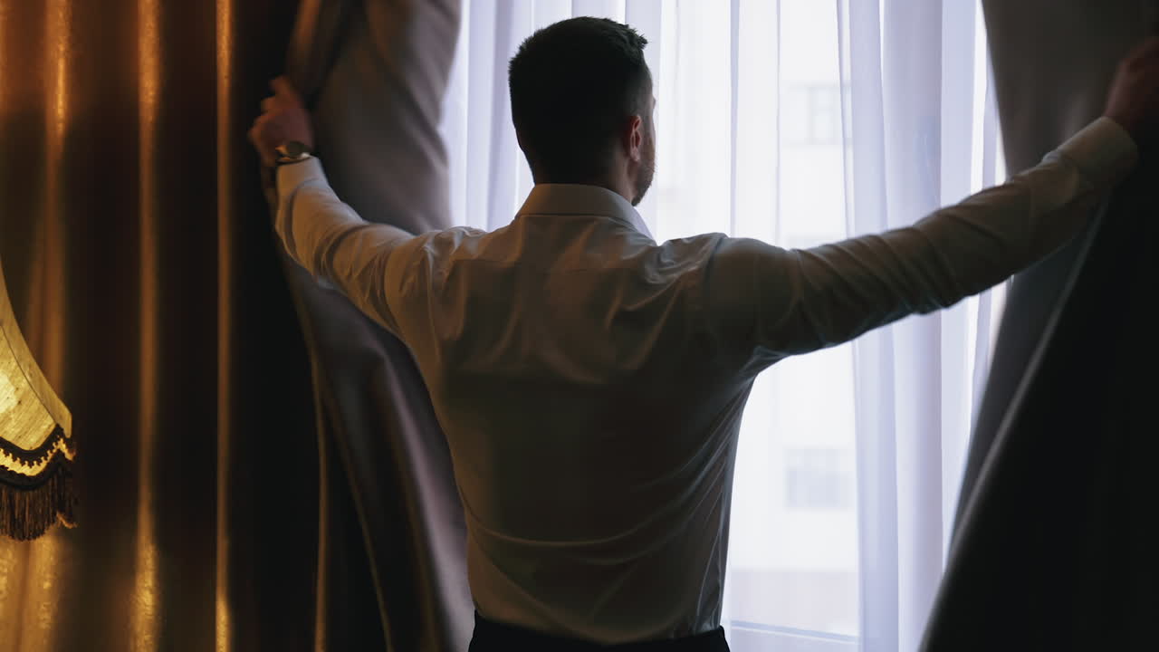 Young man opening curtains at hotel room. Man in white shirt opening the dark window curtains in a room. Relaxed man looking on the window. Back view.