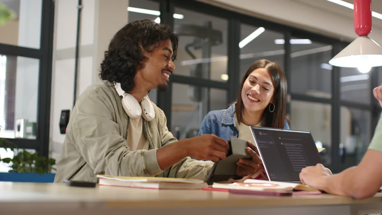 Diverse peers studying at table, man flipping tablet sparking chat, woman glancing laptop, smiling