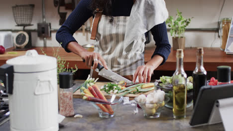 midsection of biracial woman chopping vegetables using tablet in kitchen, slow motion