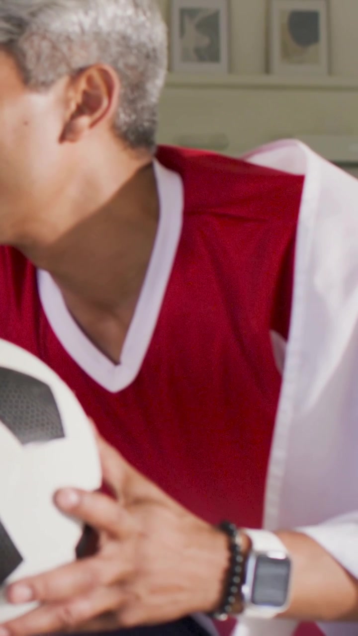 Video verticale: Uomo in maglietta rossa che reagisce, tenendo un pallone da calcio sul divano, mentre un amico indica e guarda la partita.