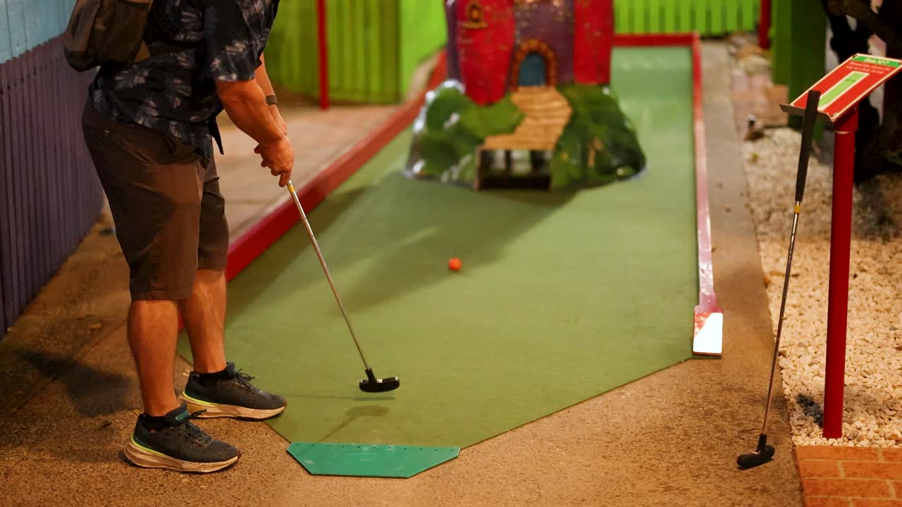 A person attempts a putt on an indoor mini golf course with colorful obstacles and ambient lighting
