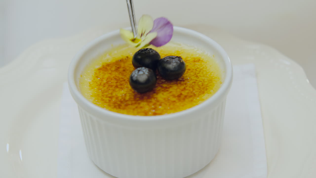 Close-up of a creme brulee with a caramelized crust adorned with blueberries and a flower, ready to be enjoyed