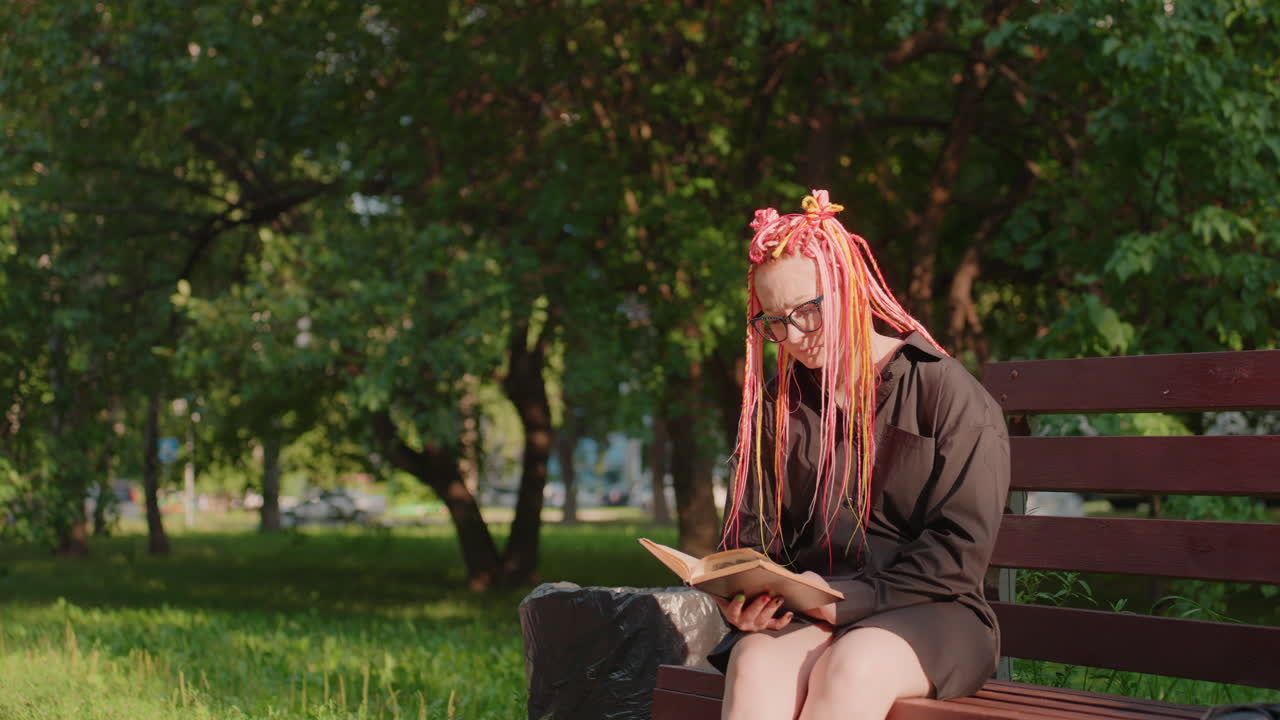 햇빛이 잘 드는 공원의 벤치에서 분홍색 땋은 머리를 하고 안경과 어두운 색 재킷을 입은 백인 여성이 가방을 옆에 둔 채 책을 읽고 있어요. 차분하면서도 어딘가 우울한 분위기 속에서 페이지를 천천히 넘기며 조용히 혼자만의 시간을 보내는 모습에서 사색적인 분위기가 느껴집니다.