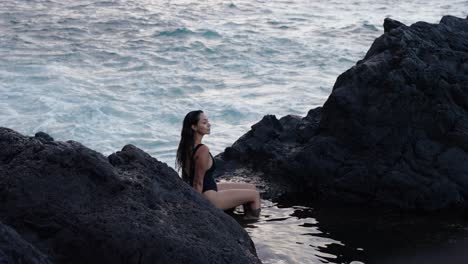woman sits quietly between volcanic rocks, gazing at the atlantic ocean in seixal, madeira during golden hour.