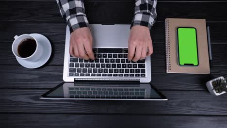 top view of the hands of a girl who type, writes emails on an laptop. near the laptop is a smartphone with a green screen and a chrome key. place for your advertising content. slow motion. close up