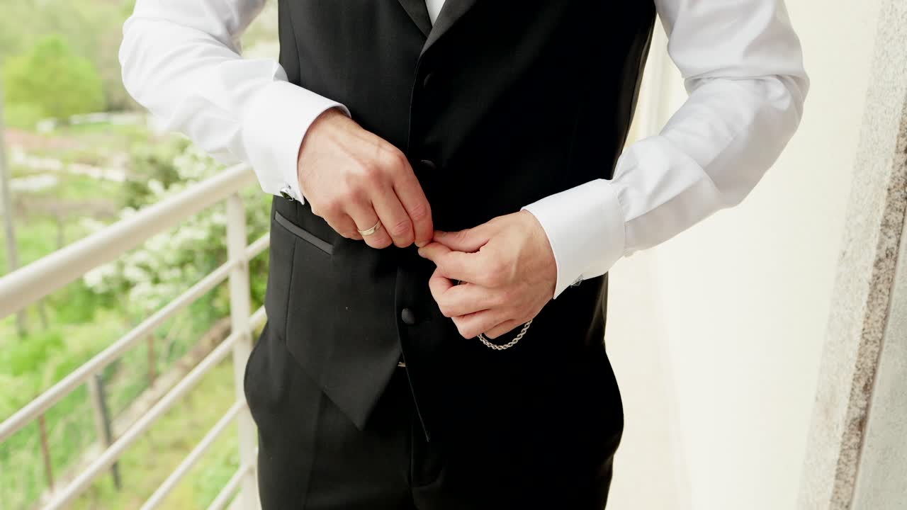 close up of groom buttoning black waistcoat with silver chain on balcony before wedding