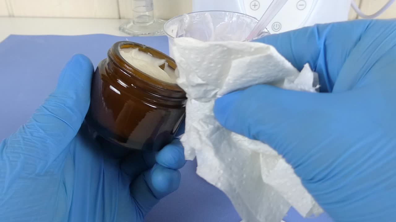 Macro view of a cosmetic laboratory scene where a gloved operator uses a spatula to carefully package a white emulsion.