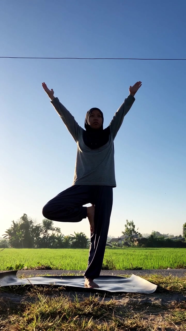 Clip al rallentatore di una donna musulmana con hijab che esegue la posizione dell'albero yoga accanto a una risaia verde all'alba. Ideale per concetti di fitness all'aperto, consapevolezza e stile di vita sano