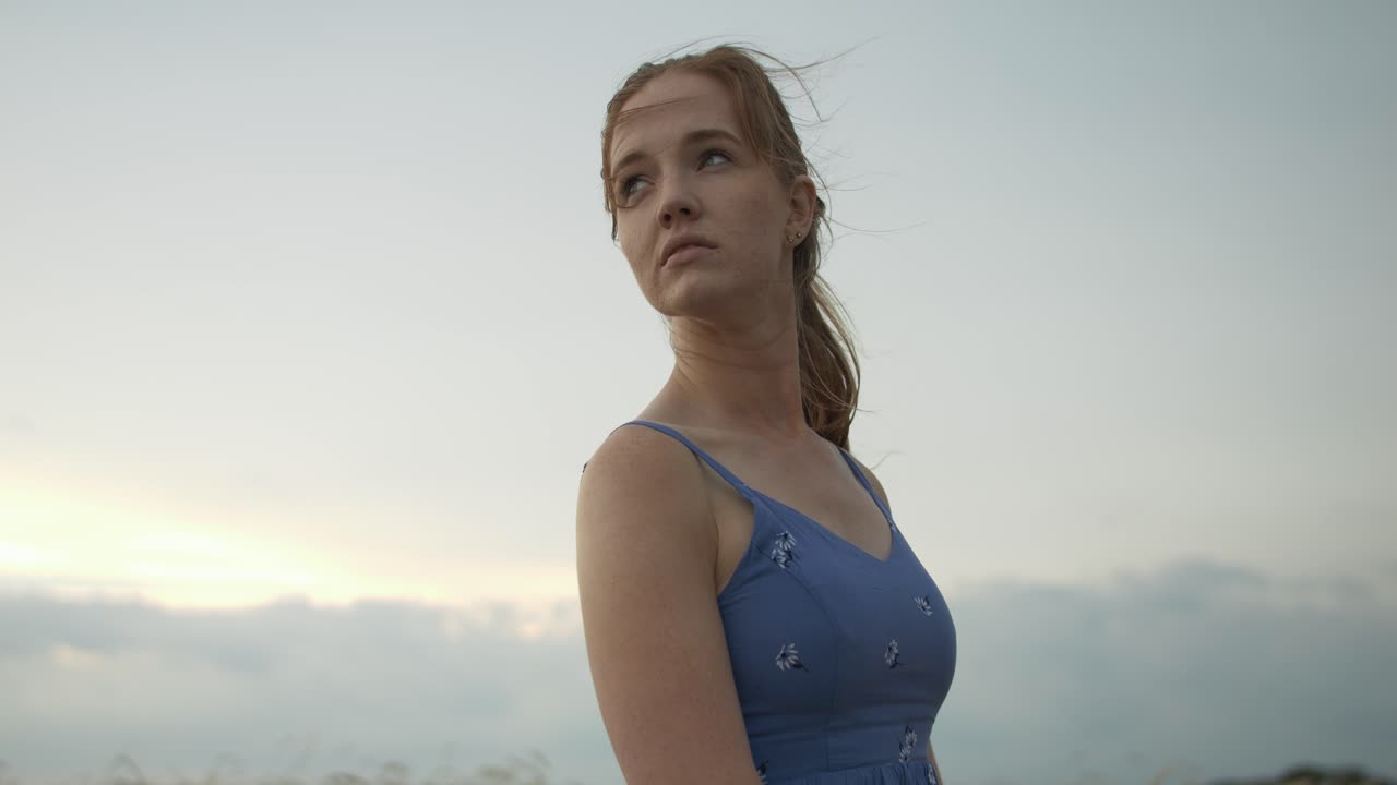 A young woman wearing a blue dress standing outside under storm clouds, moody sky in summer with hair blowing in the wind.