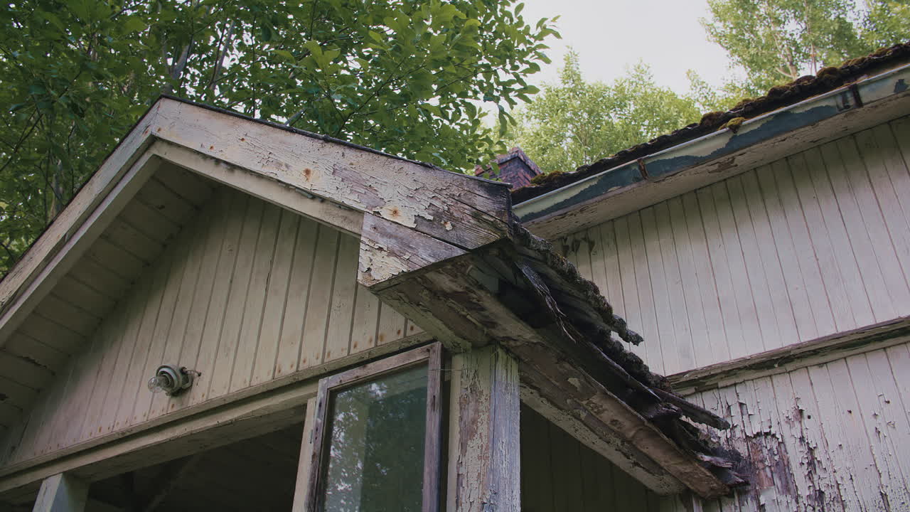 Aged house facade showing peeling paint, decayed wooden structure, and lush greenery.