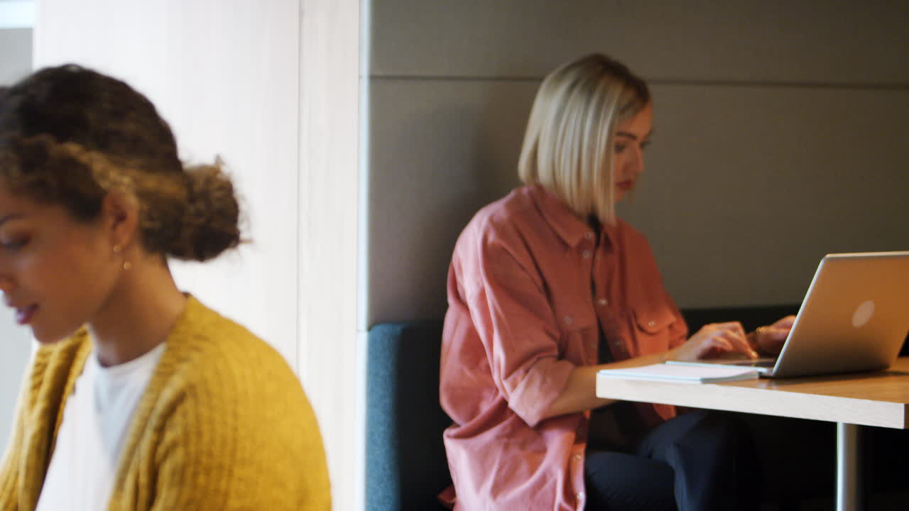 Due creative millennial che lavorano ai computer sedute in un ufficio, con messa a fuoco a passaggio dallo sfondo al primo piano
