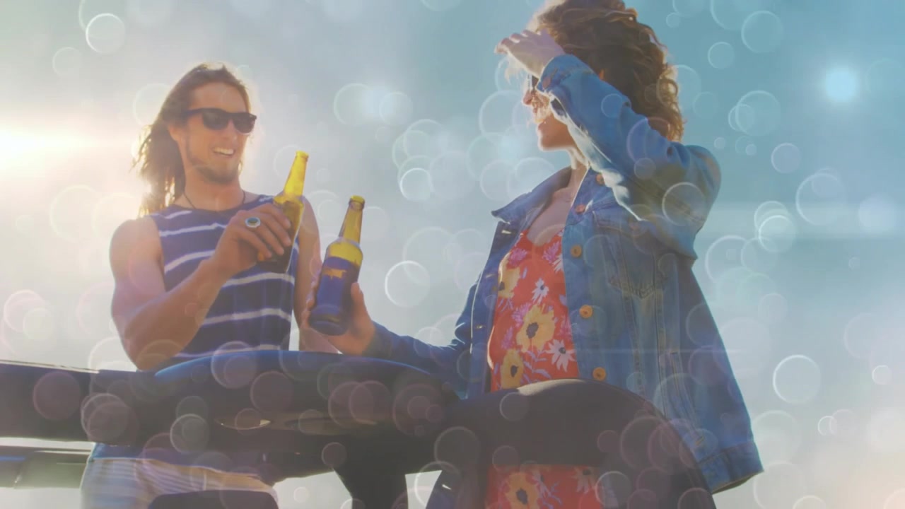 Man handing bottle, woman clinking bottles at coast rail, lens flare over faces for travel toast