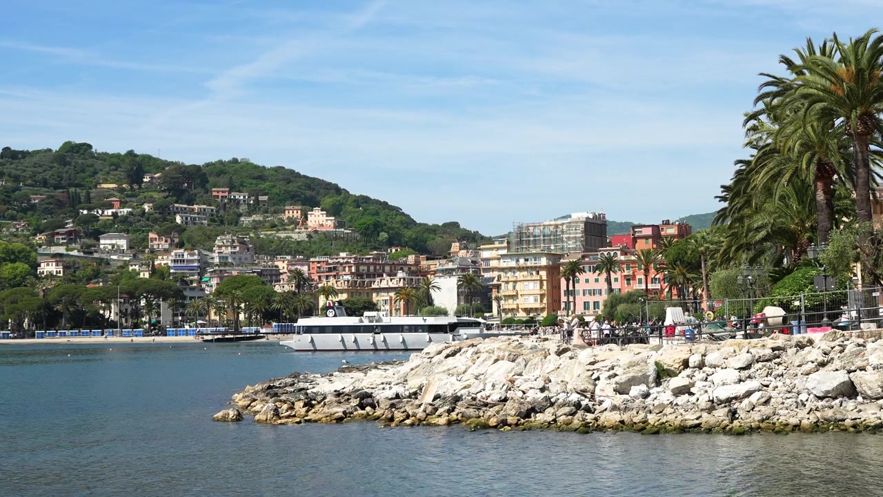 Scena costiera vivace con lungomare fiancheggiato da palme, edifici color pastello, acque calme e blu, una barca ormeggiata, colline verdi e rocce sotto una luce solare intensa, ripresa con un'inquadratura grandangolare stabile.