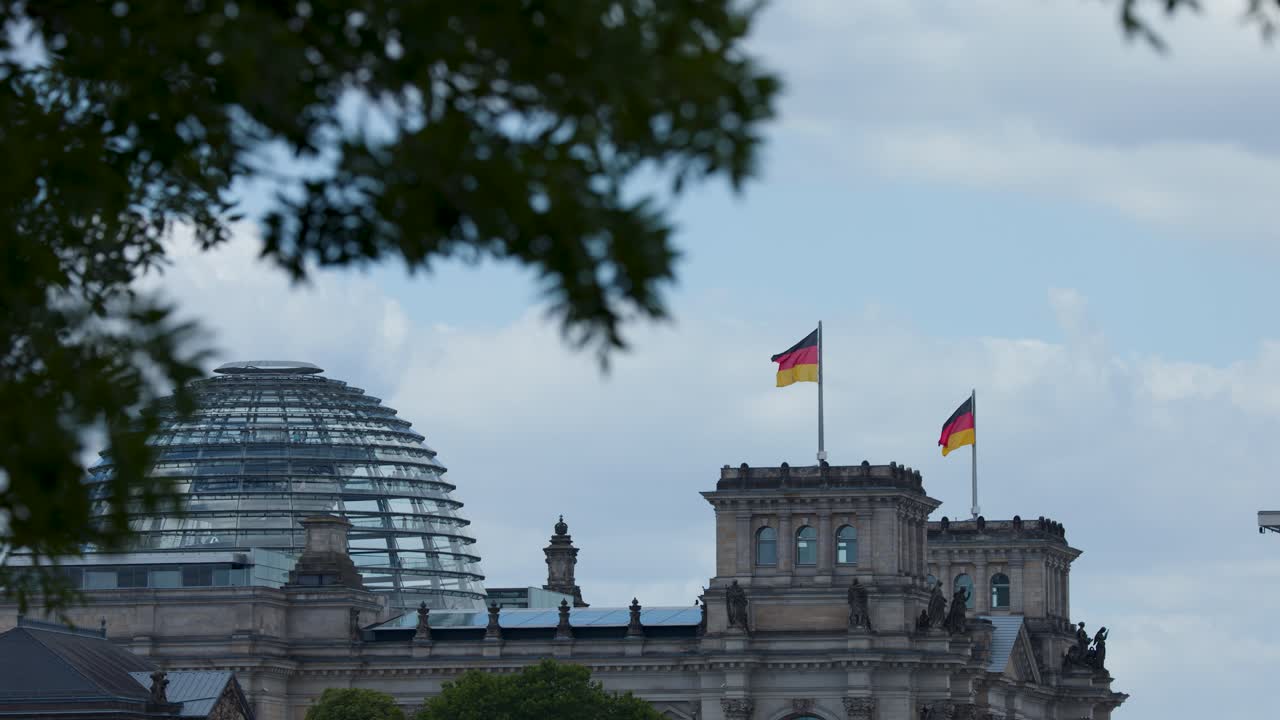 German flags wave atop historic building, glass dome visible, leafy branches, overcast daylight, static shot
