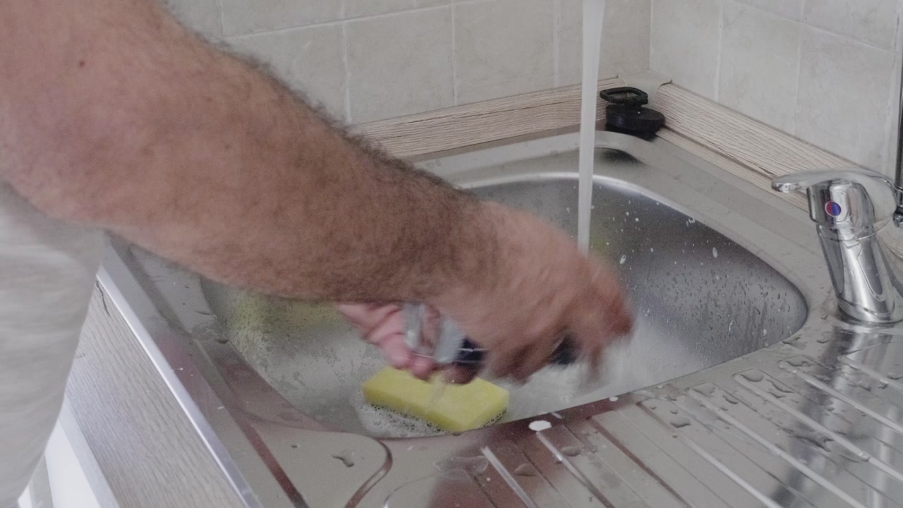 Man washing glass in stainless steel kitchen sink with yellow sponge, closeup on hands. Soap and running water domestic scene. Home hygiene and dishwashing routine.