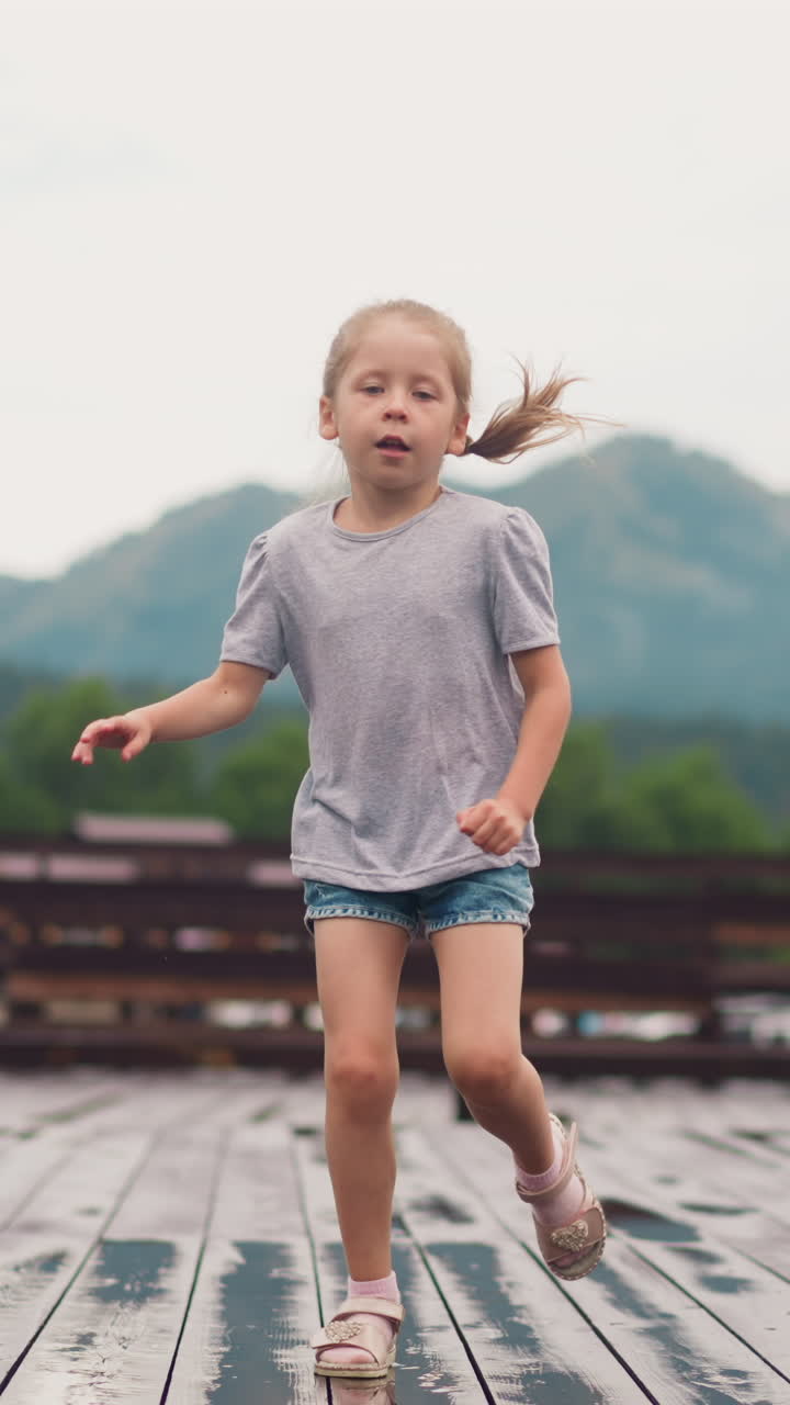 Active little blonde child in t-shirt shorts and sandals runs jumping on wet wooden board deck with small puddles after spring rain slow motion