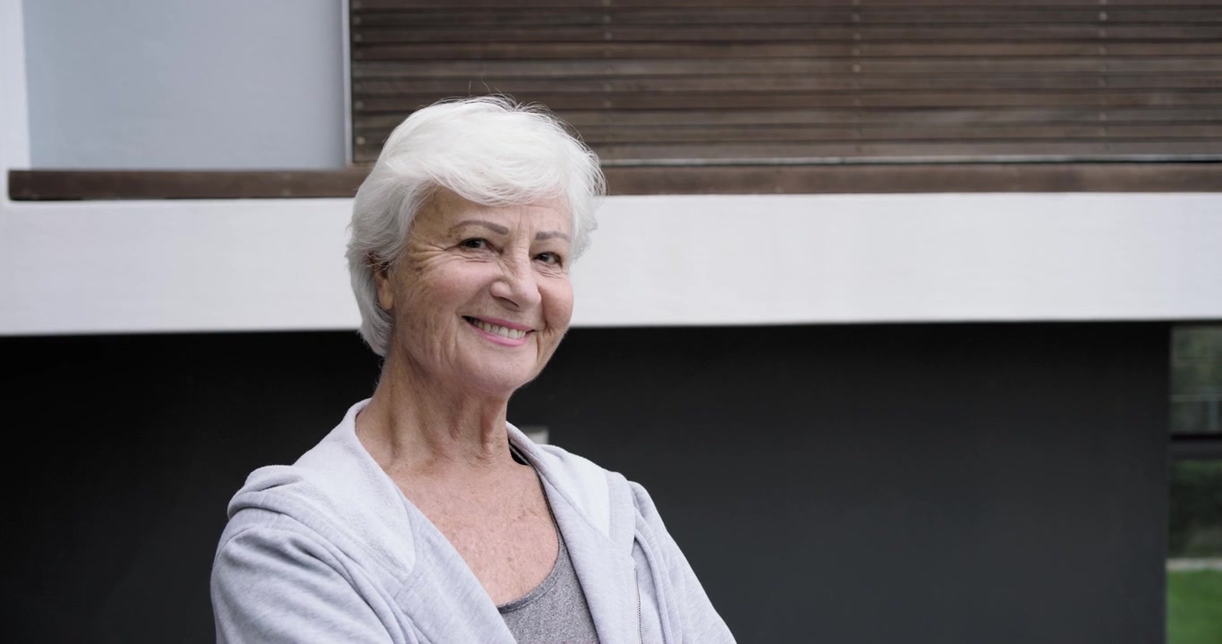 Senhora de pé na varanda, câmera se aproximando, virando a cabeça, sorrindo, com espaço para texto.