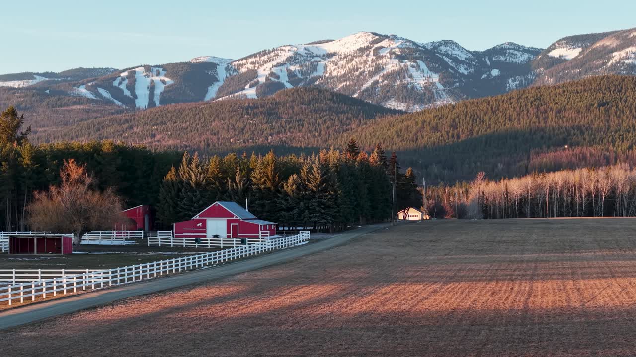 Flygbild över en lantlig gård med en röd lada och ett öppet fält nära Whitefish, Montana, med snötäckta Klippiga bergen i bakgrunden.