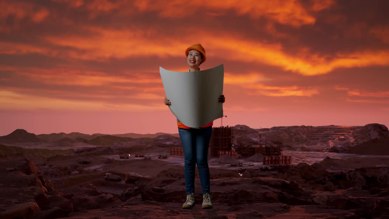 Ganzkörperaufnahme einer asiatischen Ingenieurin mit Schutzhelm, die einen Bauplan in den Händen hält und sich auf einer Baustelle mit Bergbaumaschinen umschaut