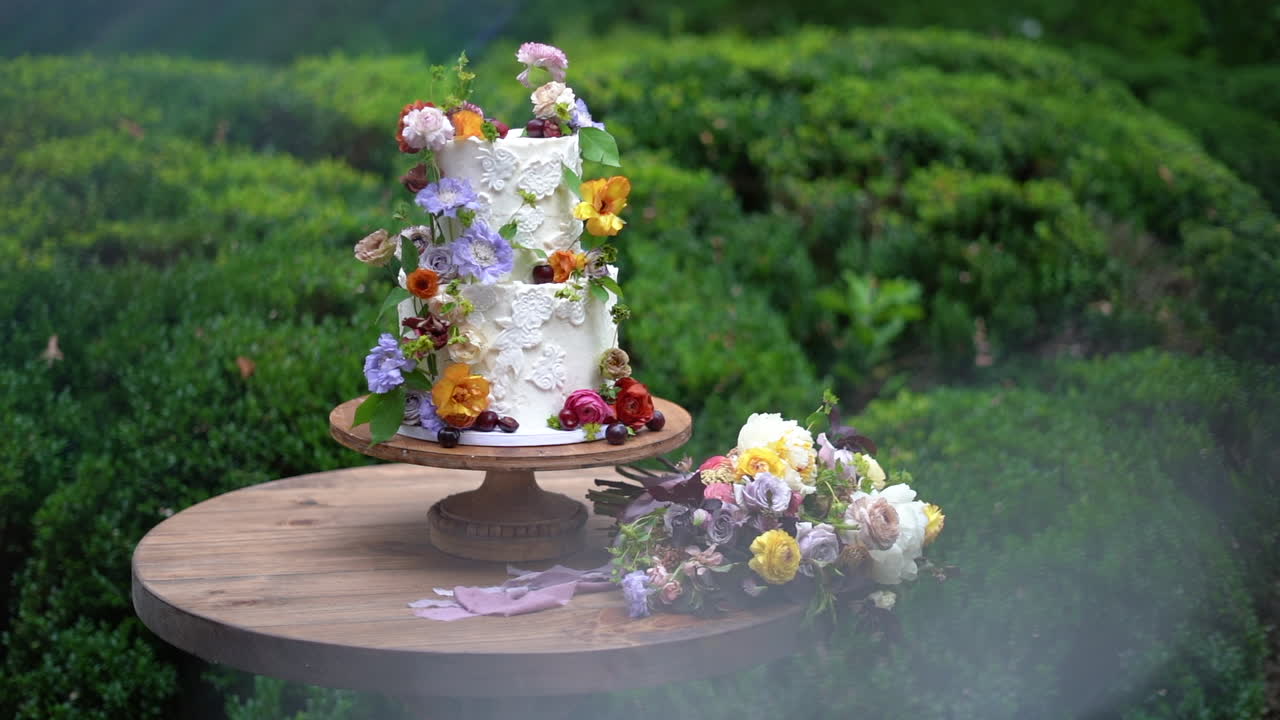 Thiết kế bánh cưới màu trắng hợp thời trang với những bông hoa đầy màu sắc và một bó hoa phù hợp trong vườn.