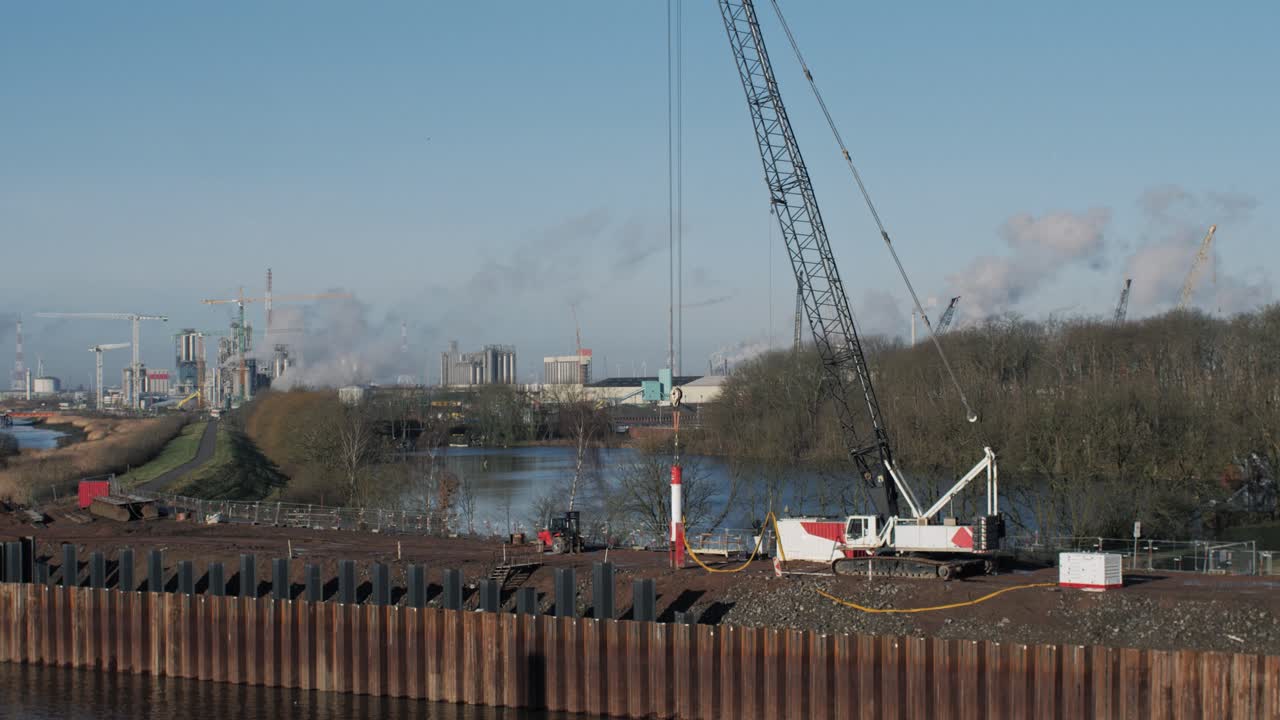 Wide shot of an industrial construction site with a large crawler crane near a river, smokestacks, and ongoing land formation work