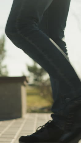 a close-up leg view of an adult walking close to an open field with trees, on a paved path, wearing jeans and black sneakers