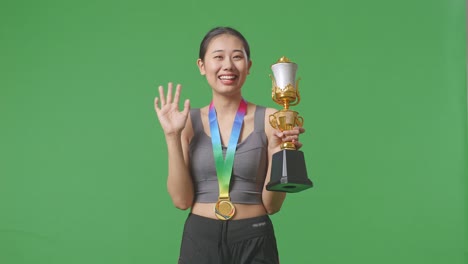 asian woman with a gold medal and trophy waving her hand and smiling to camera as the first winner on green screen background in the studio
