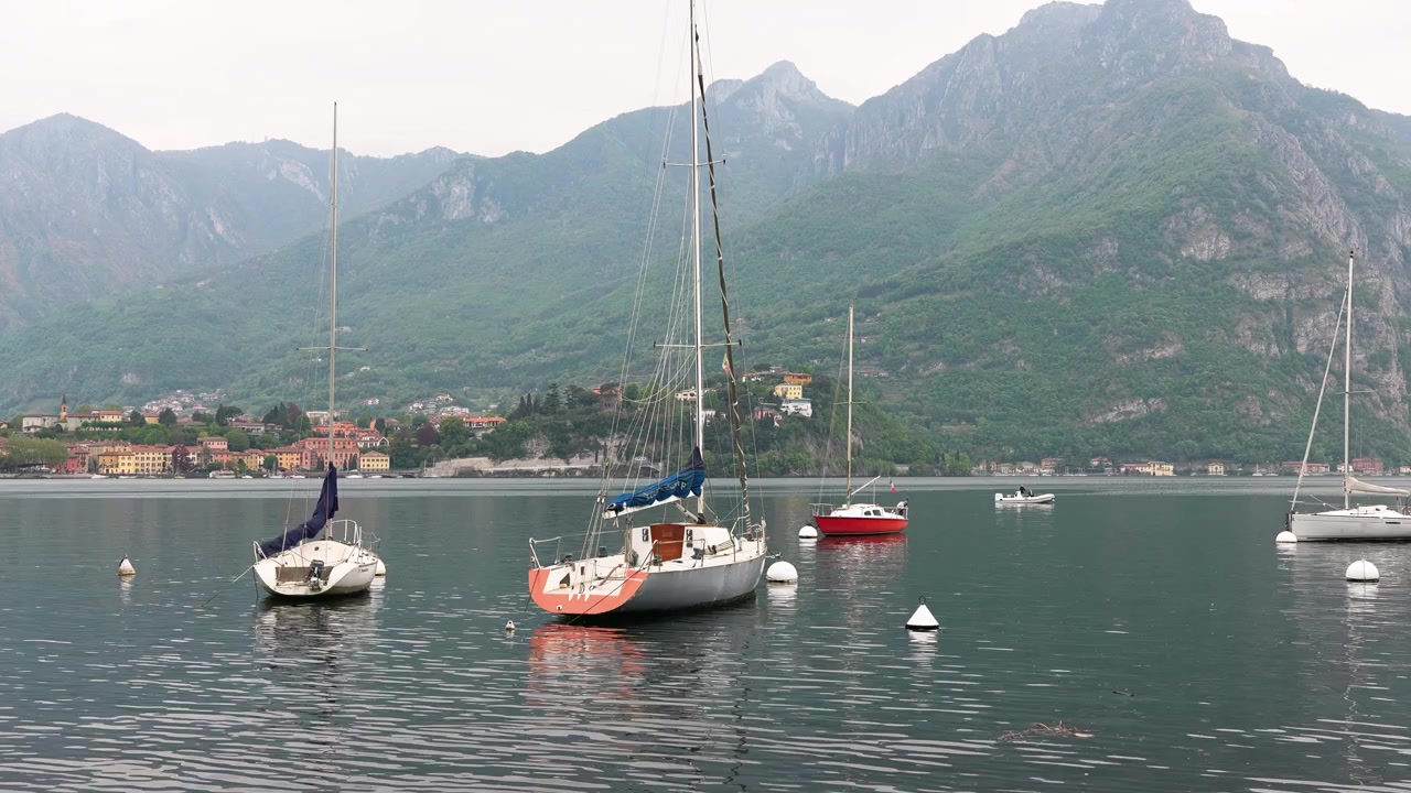 Ampia veduta panoramica di barche a vela ancorate sul tranquillo Lago di Como, vicino a Lecco, Italia, con pittoreschi edifici colorati e montagne rigogliose sotto una luce diurna soffusa, e i riflessi dell'acqua calma ben visibili.