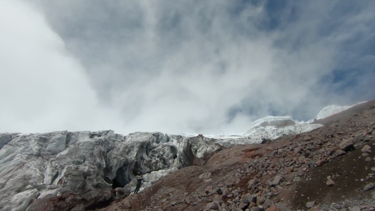 A breathtaking establishing pan of Cotopaxi volcano, Ecuador, showing rugged terrain and dramatic cloud cover with person admiring ice sheet