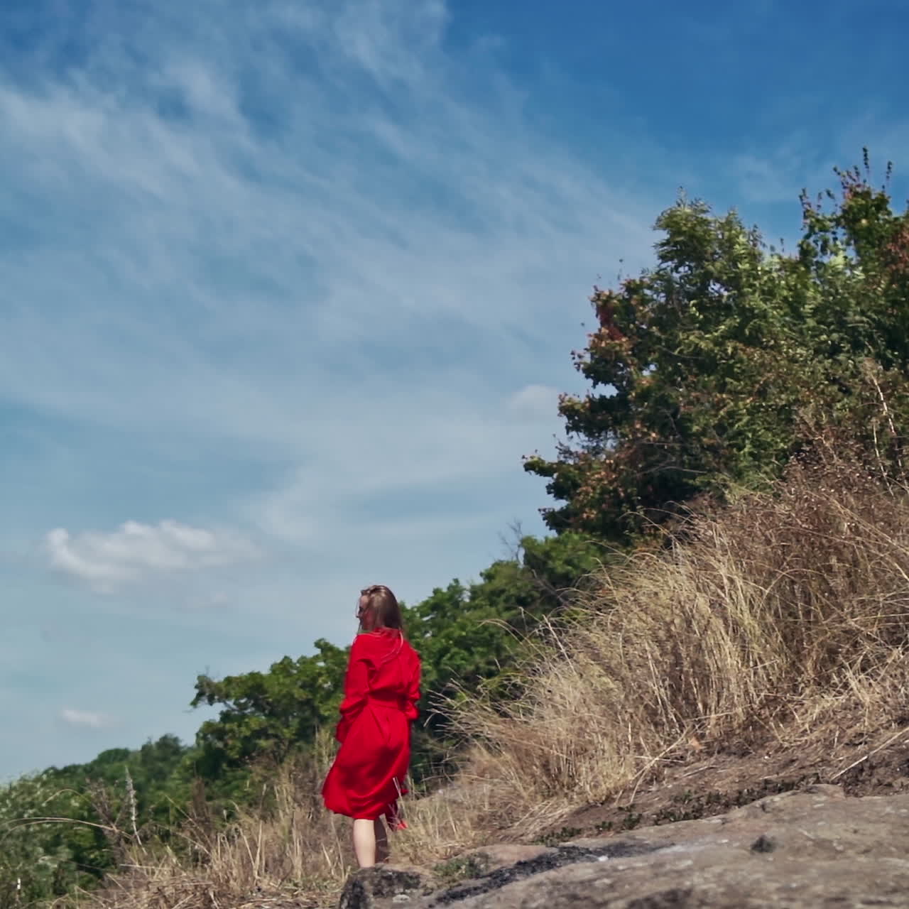 Backside view of young woman outdoors. Happy woman in beautiful red dress walking among nature. Lady enjoys summer landscape.