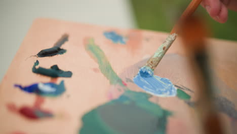 an artist s hand carefully mixes blue and white paint on a wooden palette using a brush. the close-up shot highlights the delicate process of blending colors, with various other hues visible