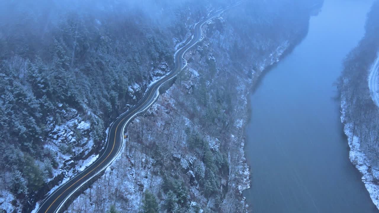 Riprese aeree di una strada panoramica innevata e tortuosa in una valle di montagna durante una tempesta di neve, con pini, un fiume, un'autostrada di montagna, scogliere rocciose e foreste durante l'inverno in una giornata fredda e azzurra.